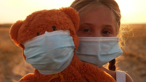child girl in medical protective mask holding a teddy bear at sunset. concept pandemic coronavirus. covid -19 girl kid face in a medical mask with a teddy bear toy. girl during coronavirus quarantine
