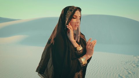 beautiful woman in black oriental abaya dress stands in desert. fashion model posing. head covered scarf hood. face is hidden by golden veil mask. Arabic nature background white sands sky sunset Dubai