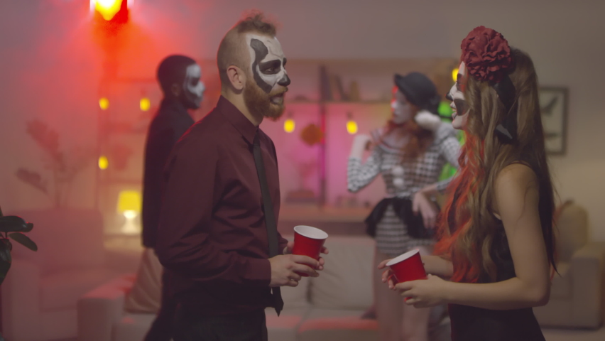 Slowmo medium shot of young man and woman in halloween costumes holding plastic cups filled with alcohol and chatting at house party. People dancing in background