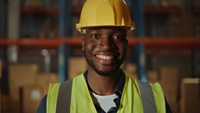 Handsome and Happy Professional Worker Wearing Safety Vest and Hard Hat Charmingly Smiling on Camera. In the Background Big Warehouse with Shelves full of Delivery Goods. Medium Close-up Portrait Royalty-Free Stock Footage #1059197429
