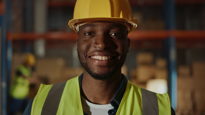 Handsome and Happy Professional Worker Wearing Safety Vest and Hard Hat Charmingly Smiling on Camera. In the Background Big Warehouse with Shelves full of Delivery Goods. Medium Close-up Portrait | Shutterstock HD Video #1059197429