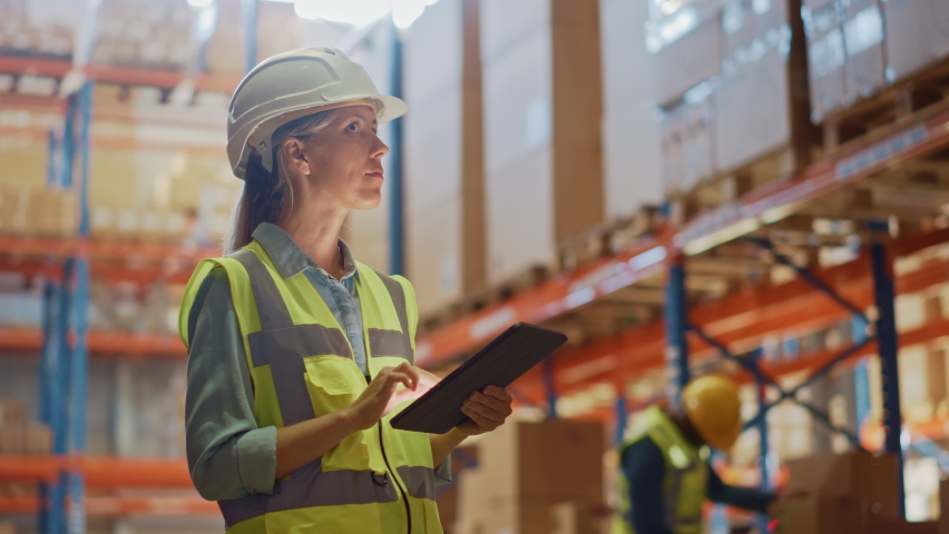 Professional Female Worker Wearing Hard Hat Checks Stock and Inventory with Digital Tablet Computer in the Retail Warehouse full of Shelves with Goods. Working in Logistics, Distribution Center Royalty-Free Stock Footage #1059197486