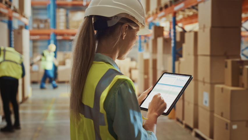 Professional Female Worker Wearing Hard Hat Uses Digital Tablet Computer with Inventory Checking Software in the Retail Warehouse full of Shelves with Goods. Delivery, Distribution Center. Side View Royalty-Free Stock Footage #1059197489