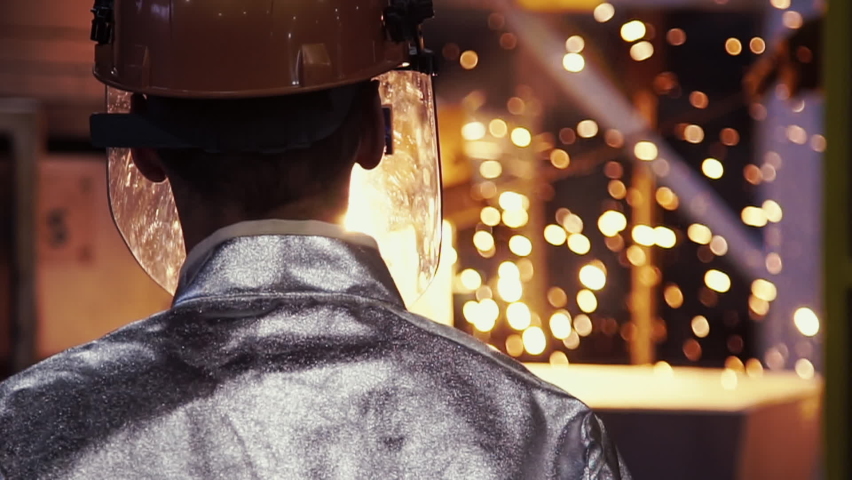 Cinematic rear view of factory worker in helmet at modern metal production plant. Foundry, metal cast, bright yellow sparks in the background, selective focus, bokeh, light. slow motion Royalty-Free Stock Footage #1059197615