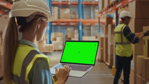 Professional Female Worker Wearing Hard Hat Holding Laptop Computer with Green Chroma Key Screen in Landscape Mode in the Retail Warehouse full of Shelves with Delivery Goods. Side view
