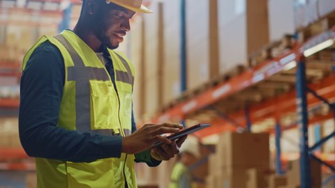 Male Worker Wearing Hard Hat Checks Products Stock and Inventory with Digital Tablet Standing in Retail Warehouse full of Shelves with Goods. Arc Shot Moves to People Operating Forklifts and Trucks
