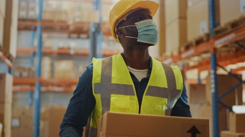 Portrait of Handsome Male Worker Wearing Medical Face Mask and Hard Hat Carries Cardboard Box Walks Through Retail Warehouse full of Shelves with Goods. Safety First Protective Workplace. Slow Motion