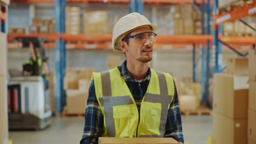 Handsome Male Worker Wearing Hard Hat Holding Cardboard Box Walking Through Retail Warehouse full of Shelves with Goods. Working in Logistics and Distribution Center. Front Following Slow Motion Shot | Shutterstock HD Video #1059197996