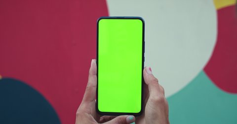 Close up view of female hands with trendy fingernails design holding smartphone and pressing on mockup screen. Concept of green screen and chroma key. Colourful spots background