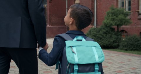 Backside view of little child with bag holding hand in hand his father in suit. Male person taking his son to school. Focus on boy. Concept of education and family