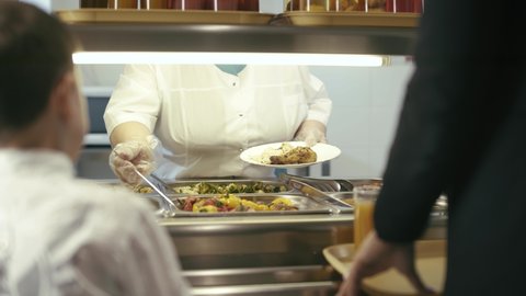 Canteen worker lays a portion stuffed peppers, rice and chicken on the plate in modern school canteen.