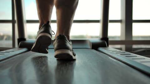 Legs of a male athlete on a treadmill. Close-up of male excellent legs with big muscles in sneakers in the gym overlooking a large window.