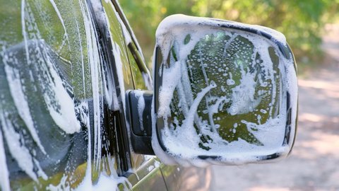 A man in a black glove washes a car mirror with a sponge. Car wash. Car in foam, car service.