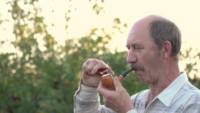 Slow-motion video of a Sad man in a shirt and with a mustache of Caucasian appearance lighting a pipe .A man lights tobacco with a lighter and lights it in the Golden hour in the garden.