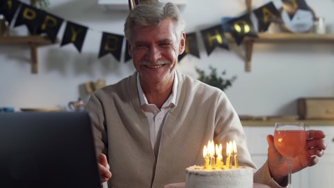 Happy smiling senior man celebrating birthday with family through online video call, receiving congratulations, blowing up candles, feeling overjoyed. Social distancing, self isolation.