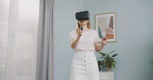 Close up of smart intelligent futuristic woman wearing virtual reality headset and holding controllers and painting at virtual display at home. 