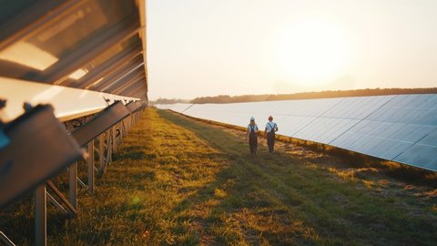 Photovoltaic field producing solar energy. Couple of workers examining solar batteries and communicating on business walking between rows. Ecological future.