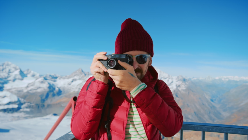 Portrait of millennial travel blogger or social media influencer, hipster man with tattoos in red puffy jacket and beanie hat make photos on analog film camera at mountain top. Wanderlust winter trip