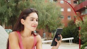 Happy young latin woman talking and texting on smart phone, wearing casual clothes sitting on bench. Generation z students life, online shopping near campus, social media and video conference.