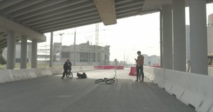 Tired bored friends with bikes standing down on street using smartphones under the bridge with street in background. Cool young bmx riders . Urban outdoors lifestyle 4k raw video footage