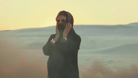 beautiful sad woman in black oriental dress abaya walks in desert. black smoke  war after explosion in background . head is covered  scarf hijab face is hidden. Arabic nature white sand dramatic sky