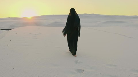 back silhouette woman in black oriental dress abaya walking in desert, rear view. head is covered scarf hijab, Long dress flying in wind. Arabic nature background, white sands, sunset sky Dubai UAE