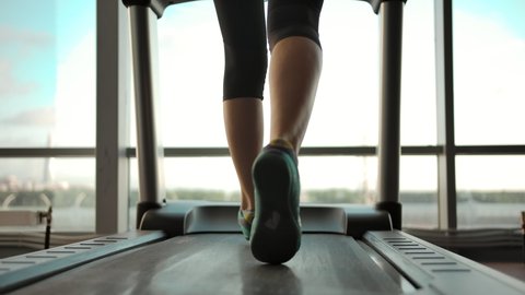 Beautiful sexy female legs on a treadmill in the gym. Vertical panning of slender cute caucasian girl blonde runs on a treadmill and looks out the window. Slow motion