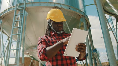 Young handsome african man, engineer with safety helmet using tablet in front of silo system. High quality 4k footage