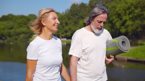 Lovely senior man and old woman holding yoga mat for exercising outdoor walking in park. Active healthy retired couple chatting and strolling by riverside after workout in summer park