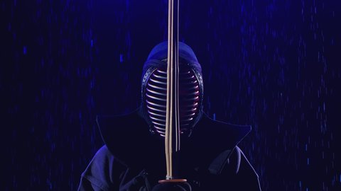 Japanese kendo. A kendo fighter stands in armor and with a bamboo sword against the background of a dark studio illuminated by blue light under the raindrops. Close up. Slow motion.