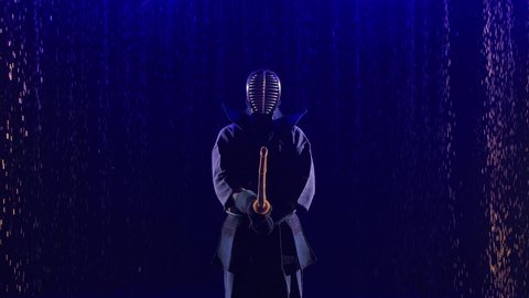 Portrait of a Japanese kendo fighter lowering his shinai towards the camera. Shot in a dark studio with blue light and rain. Close up. Slow motion.