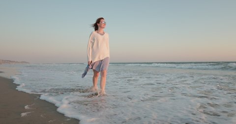Attractive female in wavy dress walking barefoot by the Pacific beach with ocean breeze blowing the hair and dress. Cinematic slow motion woman enjoying nature at sunset with blue and rose sky, 4K
