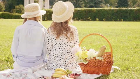 Back view.Two young beautiful hipster woman in trendy summer sundress and hats. Carefree women making picnic outside. Positive models sitting on plaid on grass, hugging and enjoying weekend 
