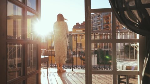 Attractive dreamy young woman in long dress going out onto terrace in her stylish apartment enjoying cozy morning vibes sunrise. Cityscape. Summertime. Inspiration.