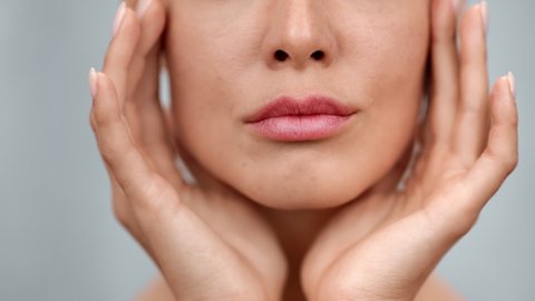 Extreme closeup feminine lips and chin applying anti aging cream on face skin. Women demonstrate moisturizing facial care touching cheek isolated on gray studio background. 4k Dragon RED camera