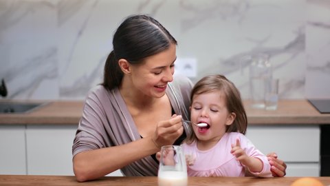 Happy young mother feeding cute daughter yogurt use spoon sitting at table in cosiness kitchen. Smiling family enjoying healthy breakfast together eating dairy food. Shot on RED Raven 4k Cinema Camera