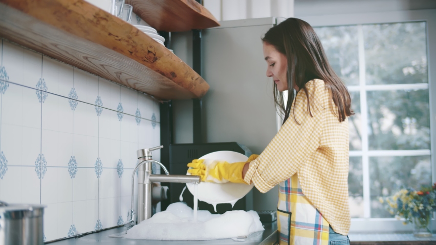 Happy attractive woman in apron and gloves washing dishes at home kitchen. Side view of pretty young housewife doing household. Beautiful female wash plates | Shutterstock HD Video #1059233144