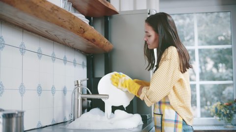 Happy attractive woman in apron and gloves washing dishes at home kitchen. Side view of pretty young housewife doing household. Beautiful female wash plates