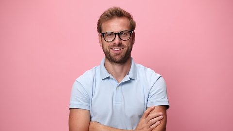 young casual guy in blue polo shirt putting glasses on, smiling, nodding and folding arms on pink background