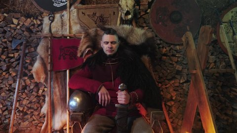 Portrait of a Viking warrior seated on a throne with a sword in his hands. In the background, spears, shields, fur cloaks, ax, firewood. A warrior dressed in medieval clothes. Camera movement