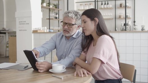 Content boss pointing at tablet screen with finger and showing something to businesswoman. Serious businessman and assistant sitting at table with cups and talking. Business and communication concept