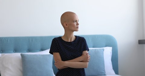 Female cancer patient sit on bed alone with arms-crossed looking out the window. Bald young woman feels afraid of upcoming operation. Struggle for life, oncology disease clinic cancer patient concept