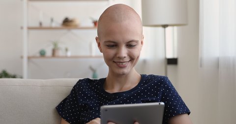 Bald young woman use tablet texting message seated on sofa, search helpful information on internet, cancer patient and recovery fulfilling life, usage of modern technology easy and comfortable concept