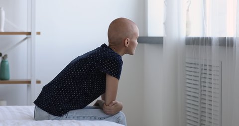 Young bald woman cancer patient sitting on bed in hospital ward alone looking out the window thinking about oncology disease feel abandoned and unhappy. Battling with tumor, recovery in clinic concept