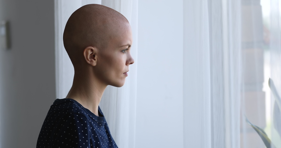 Side view cancer patient young hairless female standing in room alone near window looking outside thinking about disease feels desperate and hopeless. Oncology and rehabilitation in hospital concept | Shutterstock HD Video #1059236144