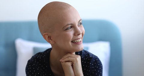Close up head shot portrait young pretty bald woman sit on bed in bedroom put chin on hands smiling looking at camera feels happy. Successful fight against cancer disease, belief in recovery concept