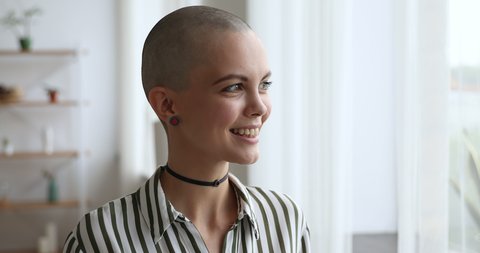 Bald gorgeous young woman wear choker striped shirt and earrings standing indoors looking out the window daydreams about career, oncology disease survivor begin new life after cancer recovery concept