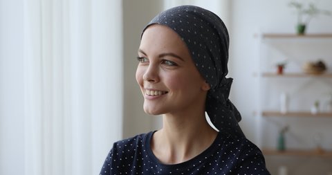 Head shot 25s young woman wear headkerchief on bald head after chemotherapy therapy smile look out the window standing indoor, welcoming day begin new life after recovery of oncology disease concept