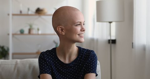 Head shot of optimistic, bald woman sitting on couch in living room smile look out the window daydreams about remission feels hopeful. Recovery over oncology disease vision into healthy future concept
