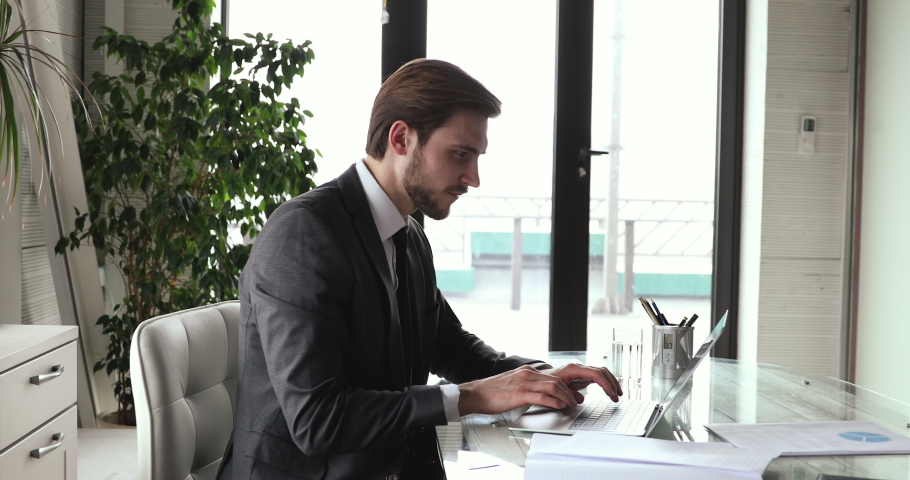 Side view focused young confident businessman in formal wear sitting at table in modern office, working on computer. Concentrated executive manager developing marketing strategy alone at workplace. Royalty-Free Stock Footage #1059240557